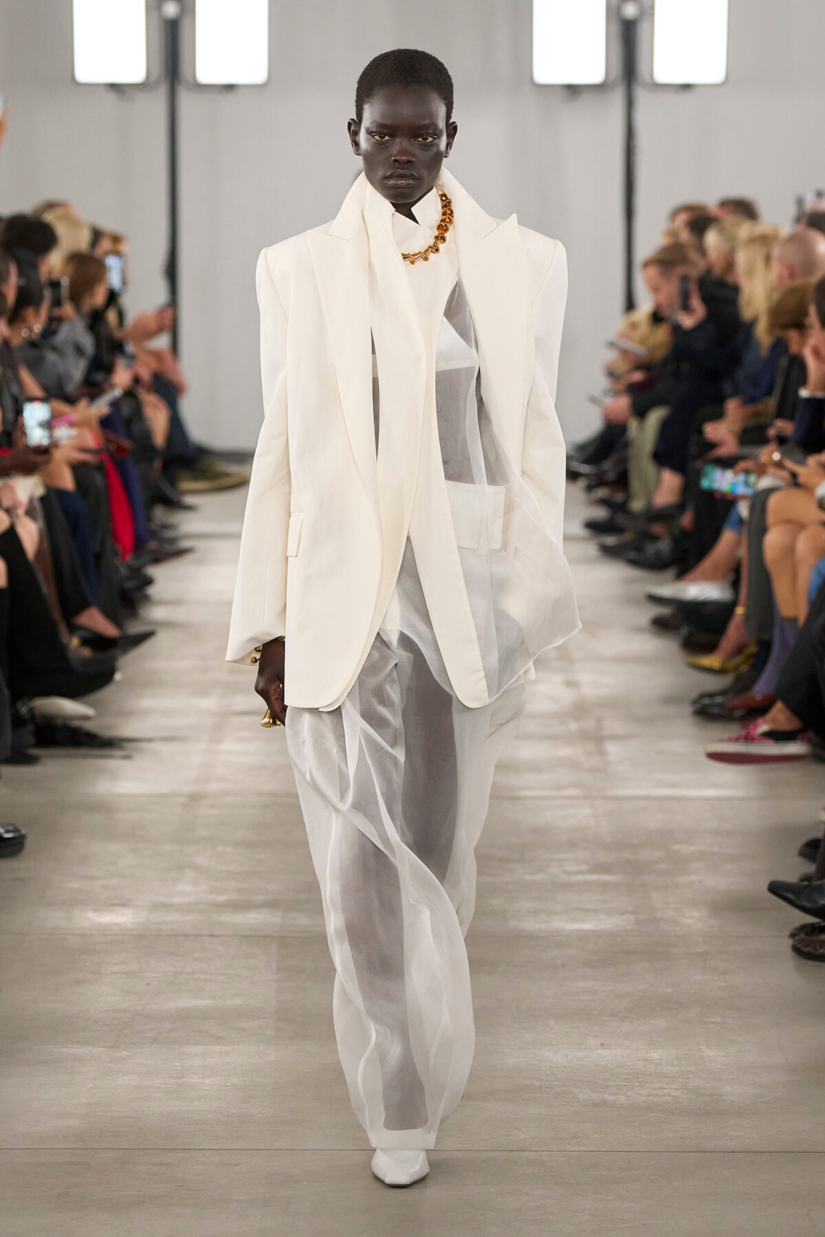Model walks a runway in an oversized white suit with a sheer inner layer and gold chain necklace, audience on both sides watching.