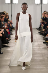 Black model walks a white, sleeveless, floor-length gown down a runway with spectators on both sides.