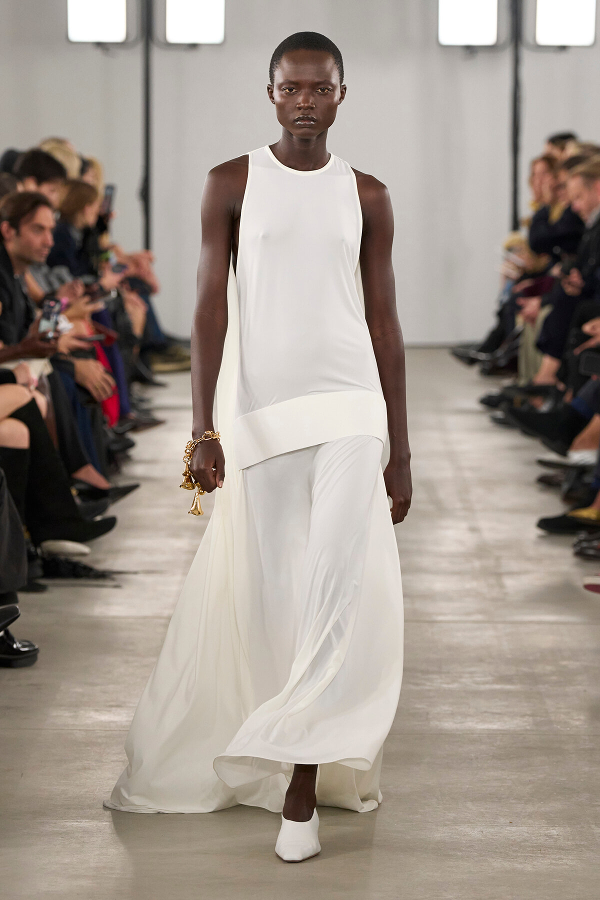Black model walks a white, sleeveless, floor-length gown down a runway with spectators on both sides.