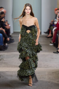 Model walking a runway in a green leopard-print multi-layered dress with thin black straps and sheer underlayer.