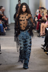 Model walks a runway in a sheer black top with bird embroidery worn over jeans with black feather patterns.