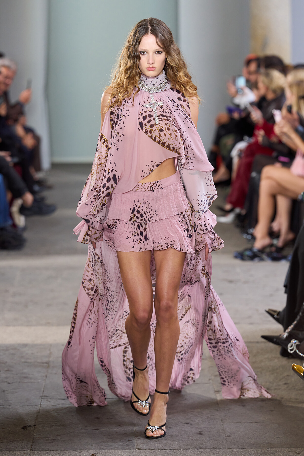Model walks a pink patterned gown with a flowing cape down the runway, audience watching on both sides.