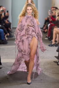 Model walks the runway in a pink, ruffled, cold-shoulder gown with leopard print and a high slit, paired with oversized chandelier earrings.