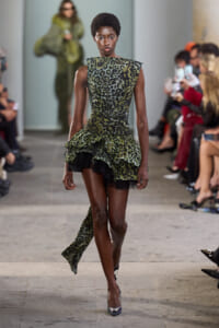 Model walks a runway wearing a green leopard-print dress with a layered ruffled skirt and matching long tail sash, black heels.