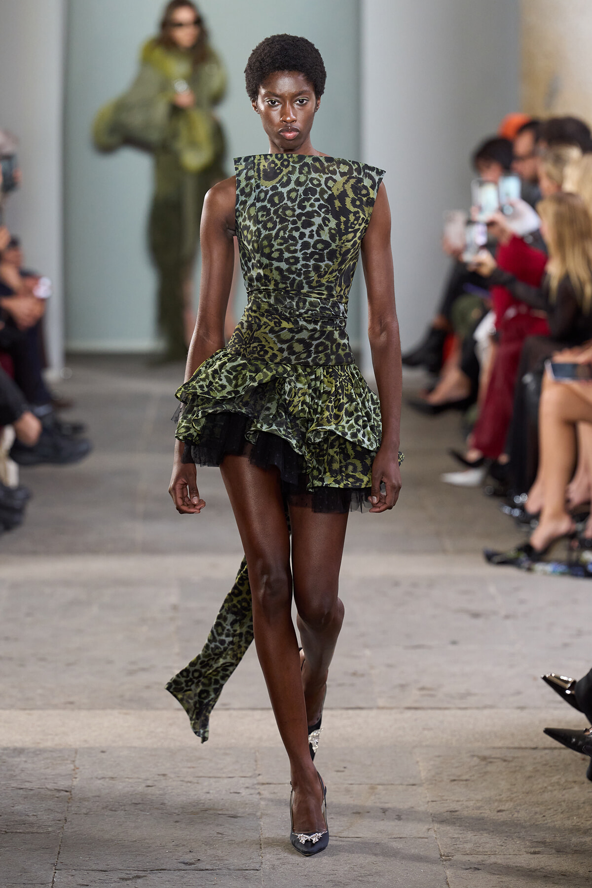 Model walks a runway wearing a green leopard-print dress with a layered ruffled skirt and matching long tail sash, black heels.
