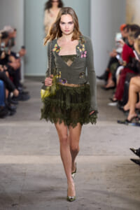 Model walking runway in olive cardigan with colorful butterfly appliqués and a dark green lace tiered skirt; audience watches in background.