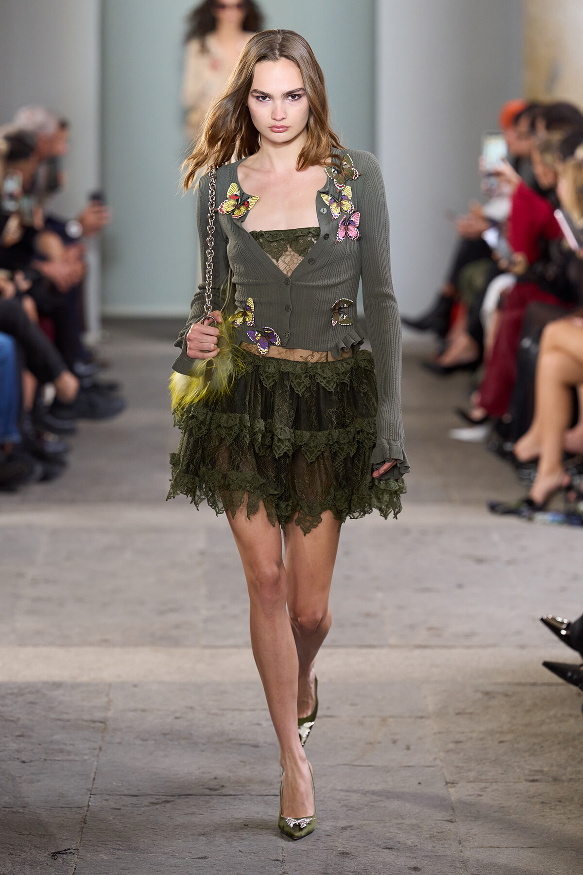 Model walking runway in olive cardigan with colorful butterfly appliqués and a dark green lace tiered skirt; audience watches in background.