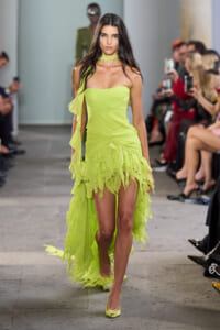 Model walks a runway in a lime-green strapless dress with a ruffled, high-low hem as the audience watches and photographs.