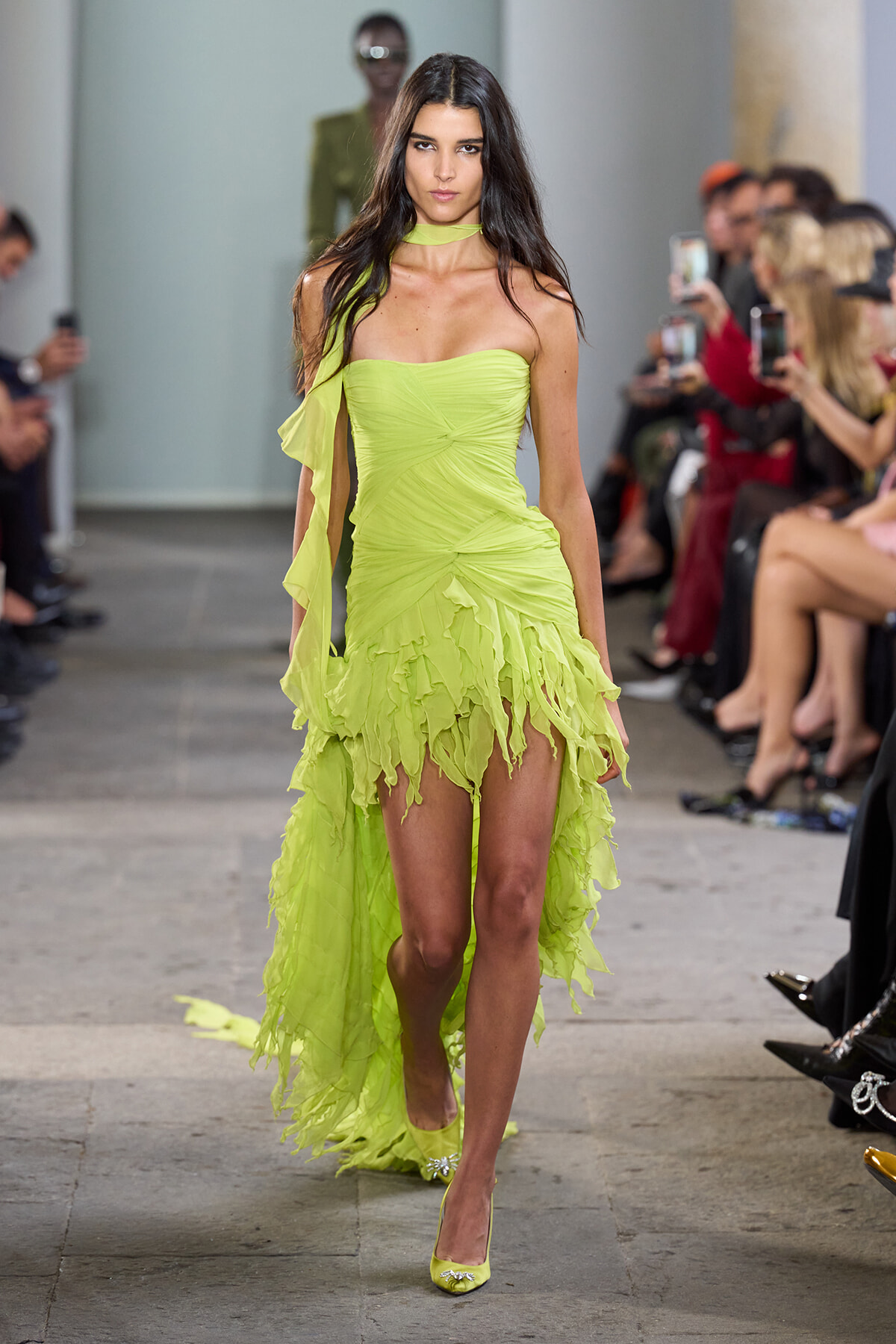 Model walks a runway in a lime-green strapless dress with a ruffled, high-low hem as the audience watches and photographs.
