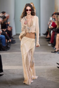 Runway model in a nude-toned ensemble with sheer cardigan, lace panel skirt, oversized sunglasses, and bold cross necklaces.