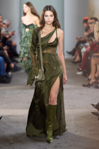 Model on a runway wearing an olive-green, asymmetrical dress with lace panels and a thigh-high slit, carrying a small crossbody bag.