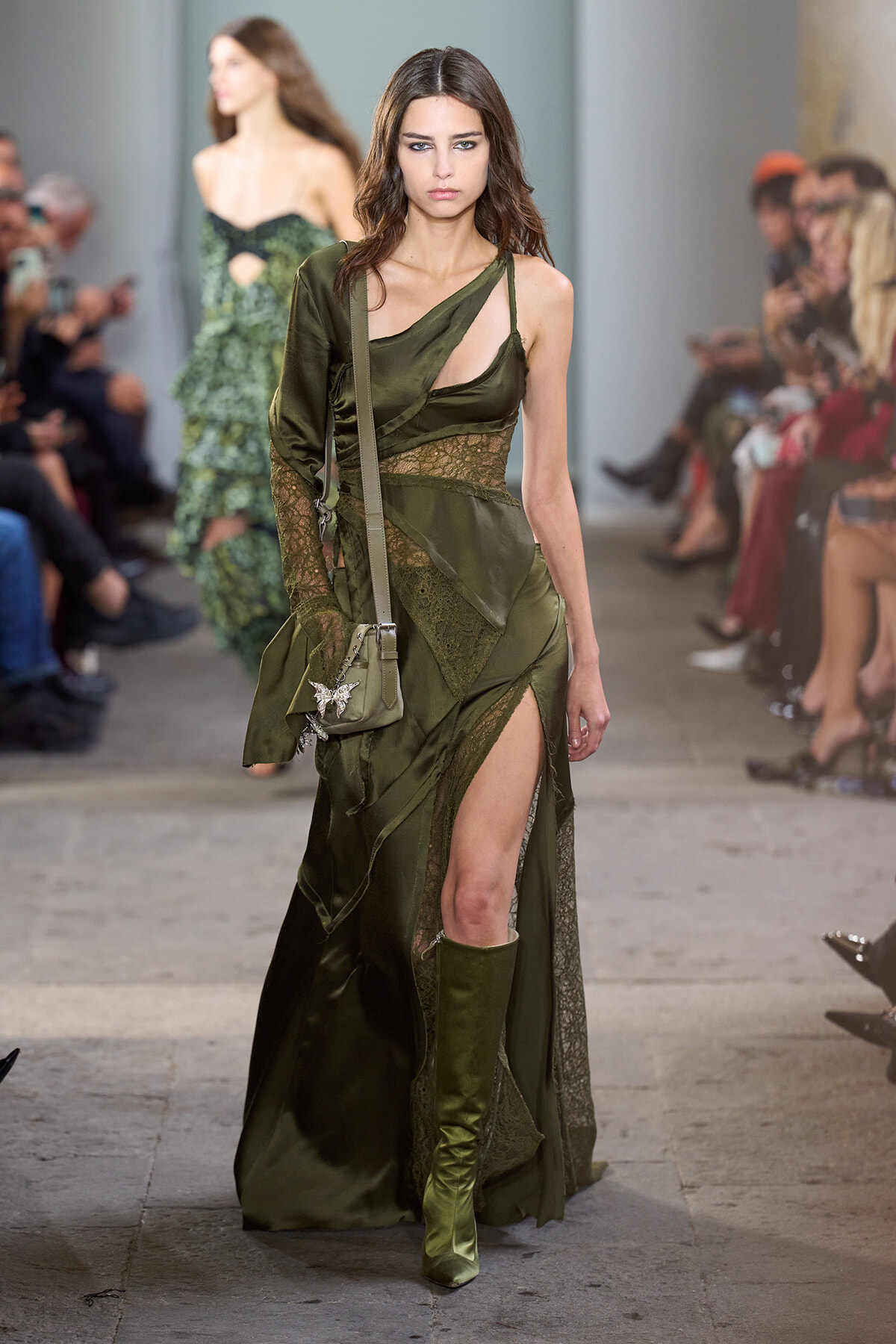 Model on a runway wearing an olive-green, asymmetrical dress with lace panels and a thigh-high slit, carrying a small crossbody bag.