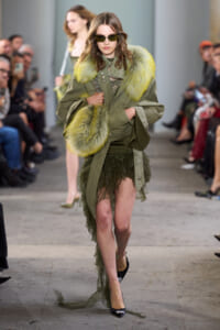 Model walks the runway in an olive-green military-inspired coat with bright lime fur trim, paired with a green lacey high-low skirt and black heels, wearing sunglasses.