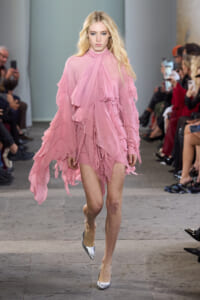 Model walking a runway in a pink, flowing, layered ruffled dress with silver heels, surrounded by spectators.