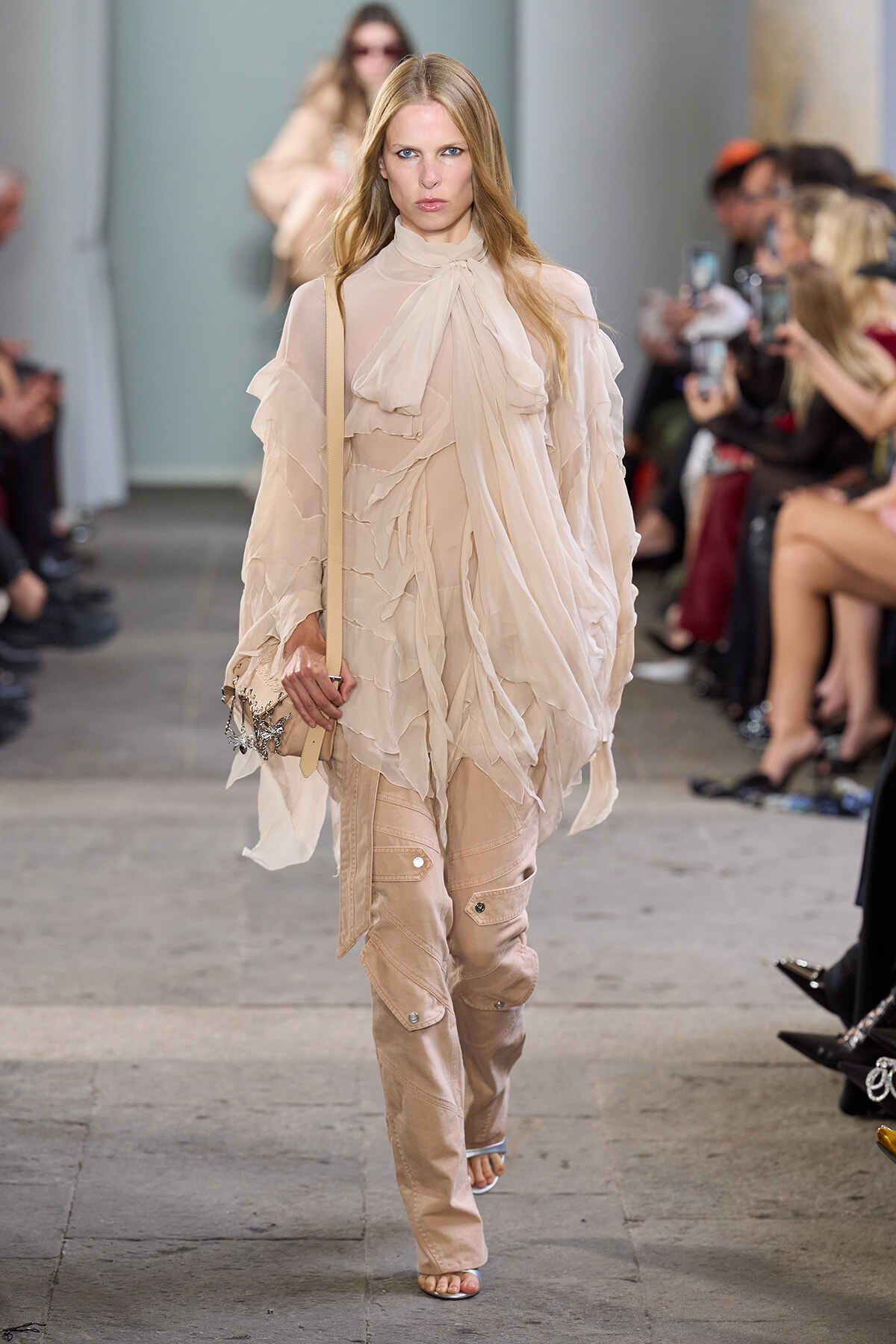 Model walking a runway in a sheer beige blouse with a draped necktie and layered ruffles, paired with light tan cargo pants and a beige crossbody bag.