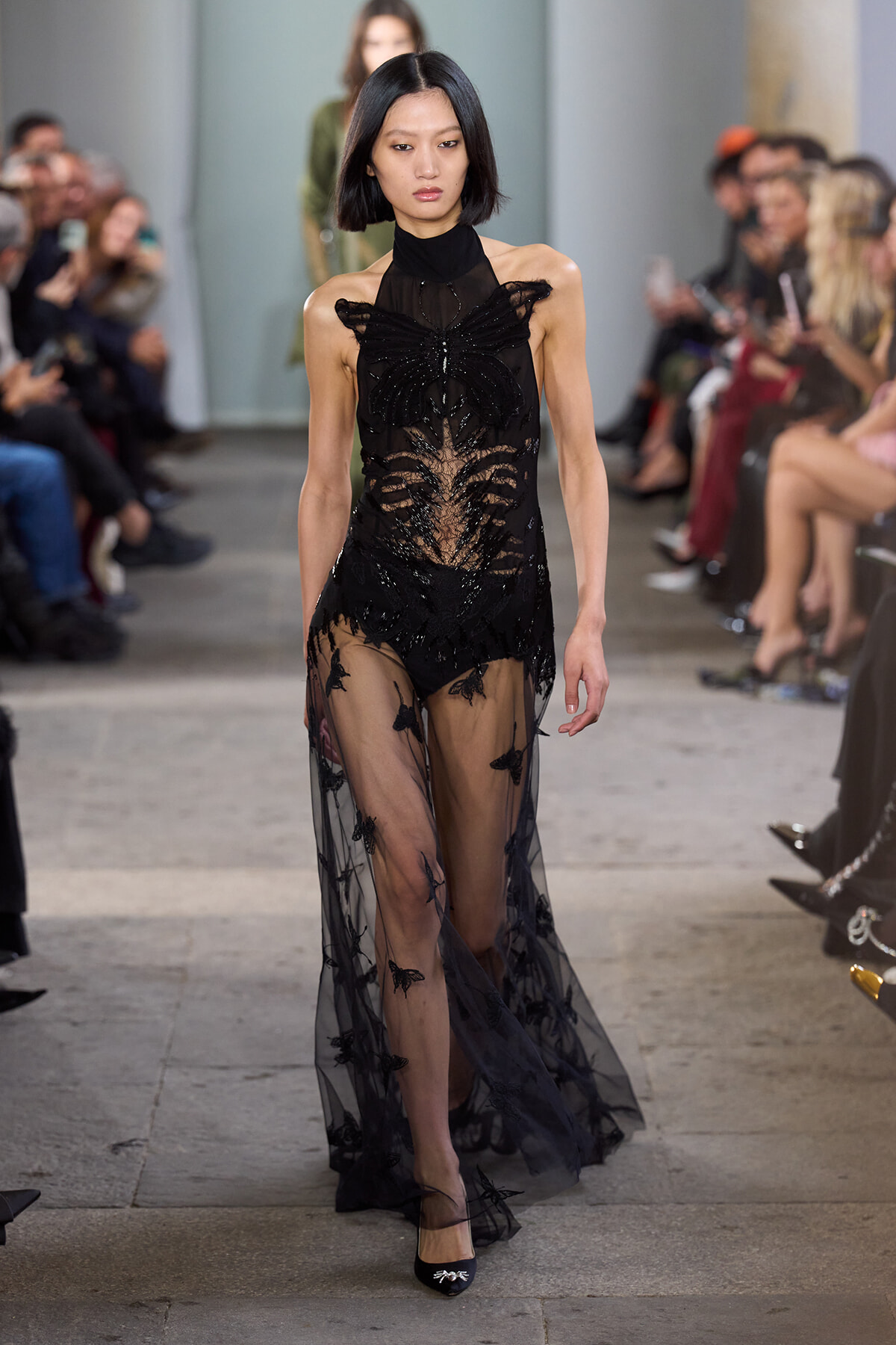 Model walking the runway in a black sheer gown with butterfly lace embellishments and a long transparent skirt, audience on both sides.