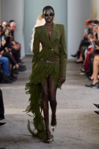 Model walking a fashion runway in an olive-green blazer and high-low ruffled skirt, wearing sunglasses and matching heels.