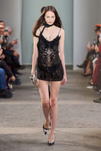 Model walking a runway in a black sheer lace mini dress with thin straps, a choker, and silver-embellished heels.
