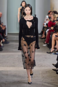 Model walks the runway in a black knit top with a deep neckline and a sheer, embroidered overlay skirt; onlookers blur in the background.