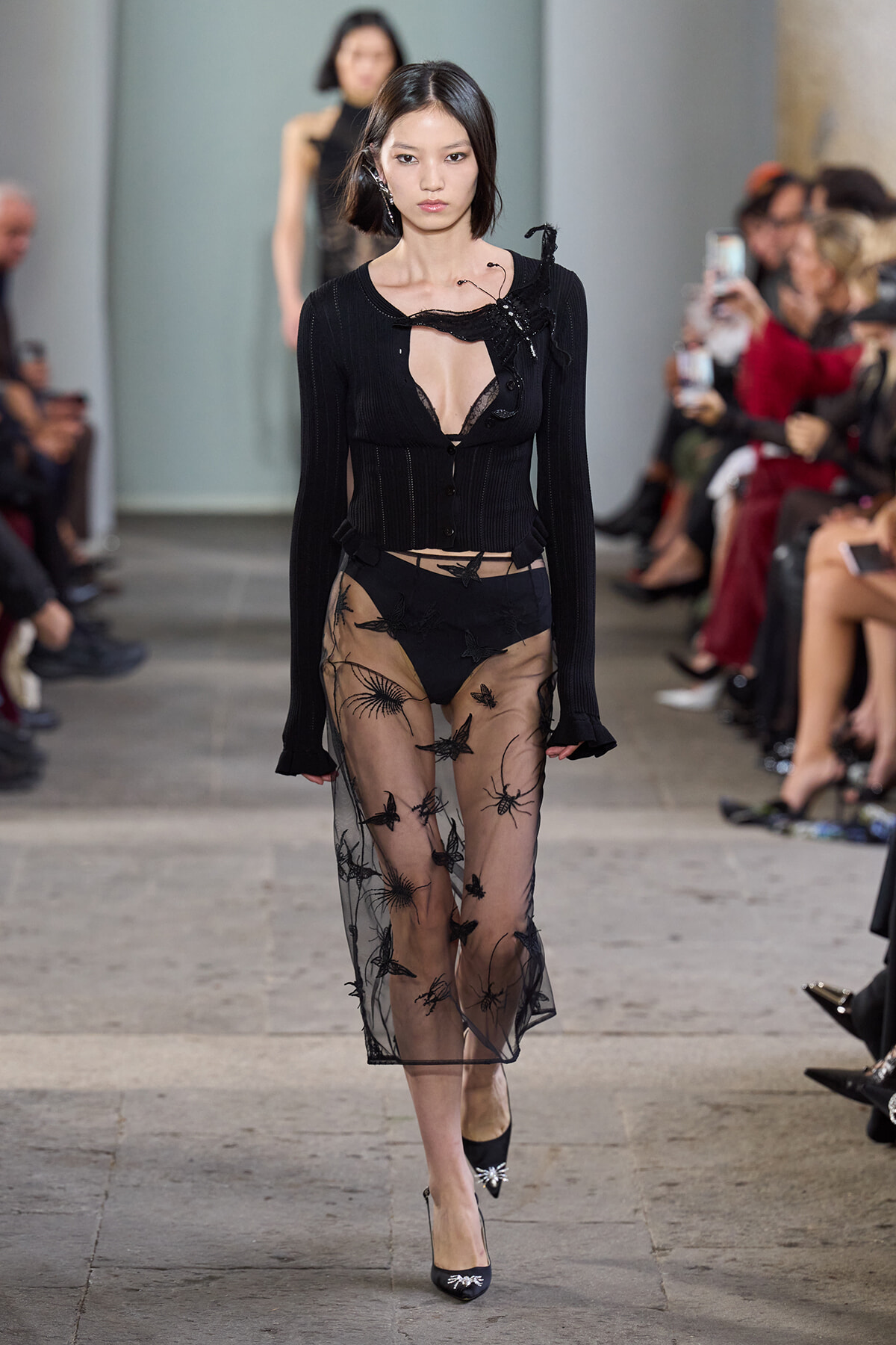Model walks the runway in a black knit top with a deep neckline and a sheer, embroidered overlay skirt; onlookers blur in the background.