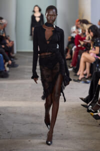 Model walks a runway in a sheer black lace skirt and cropped black top, with audience seated and watching on both sides.