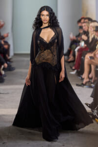 Model walking a runway in a sheer black lace gown with a cape at a fashion show, audience blurred in the background.
