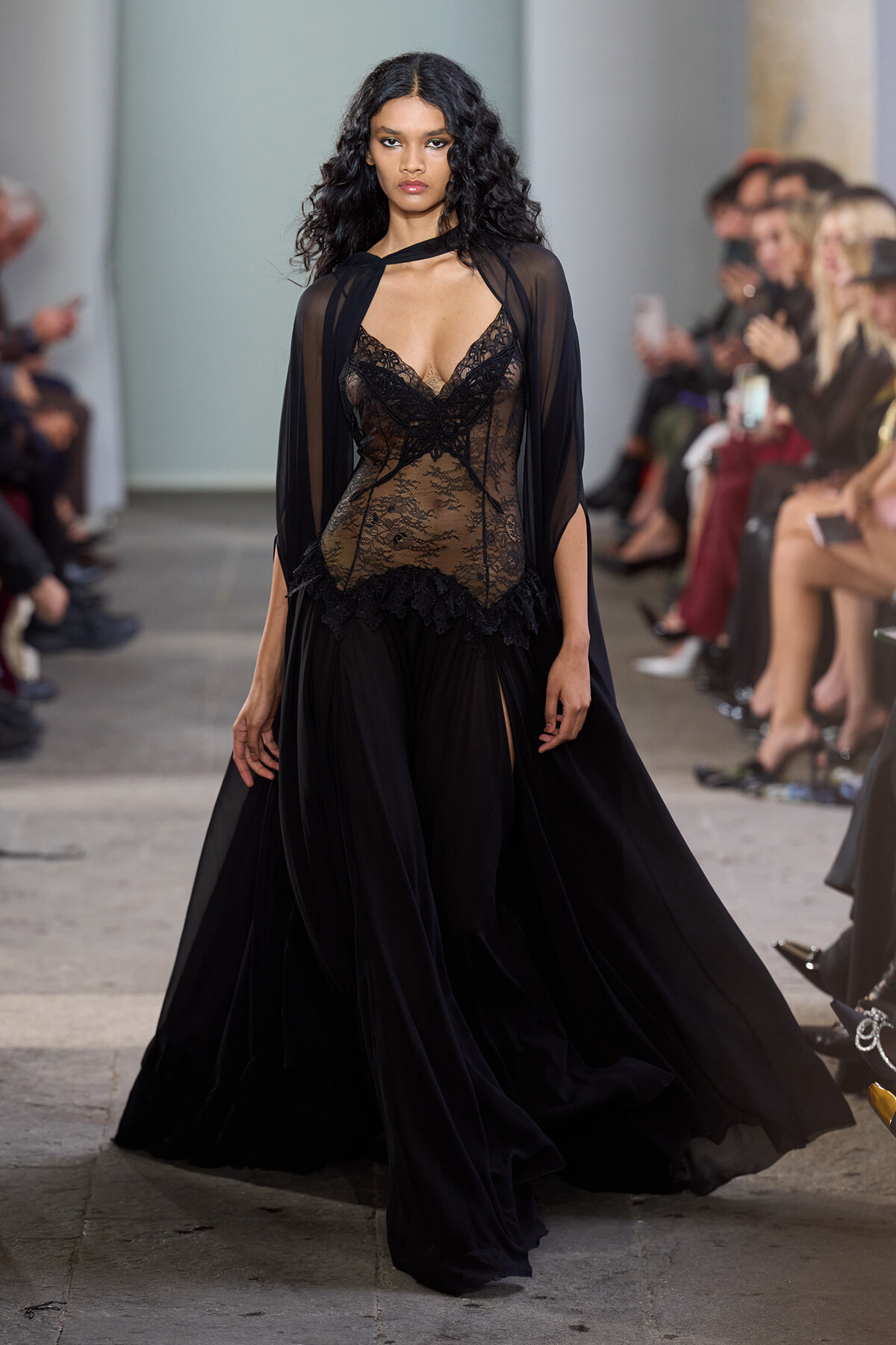 Model walking a runway in a sheer black lace gown with a cape at a fashion show, audience blurred in the background.