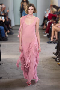 Model walks down a runway in a pale pink, sheer dress with cascading ruffles at a fashion show.