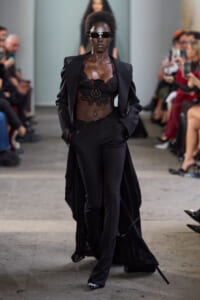 Model walking the runway in a black lace bralette, tailored pants, and a long cape-like coat with futuristic sunglasses.