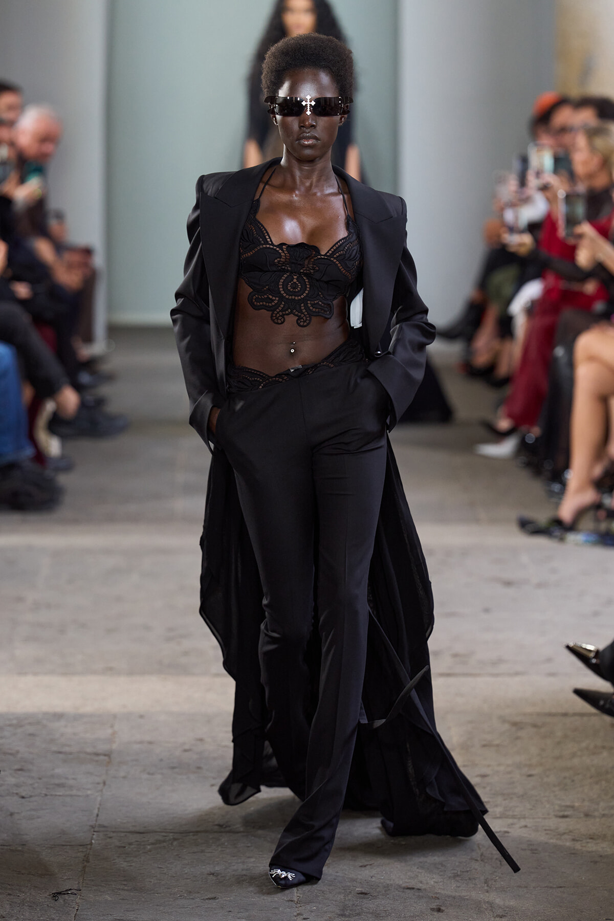 Model walking the runway in a black lace bralette, tailored pants, and a long cape-like coat with futuristic sunglasses.