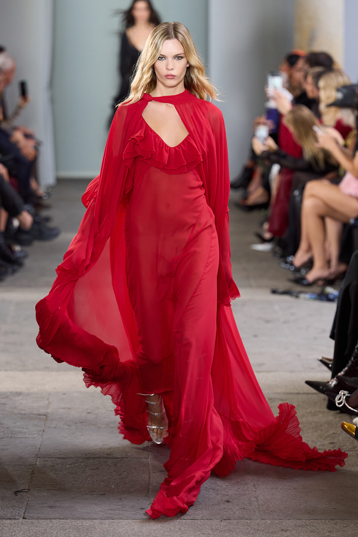 Model walks a sheer red gown with ruffles down a runway, audience watching and filming nearby.