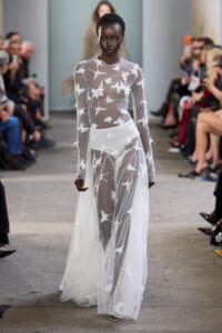 Model walks the runway in a sheer white gown covered in white star-pattern embroidery, with an audience on both sides observing the show.