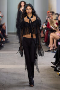 Model walks a runway in a black lace blouse with long sheer sleeves, tied scarf at the neck, and wide-leg black pants.
