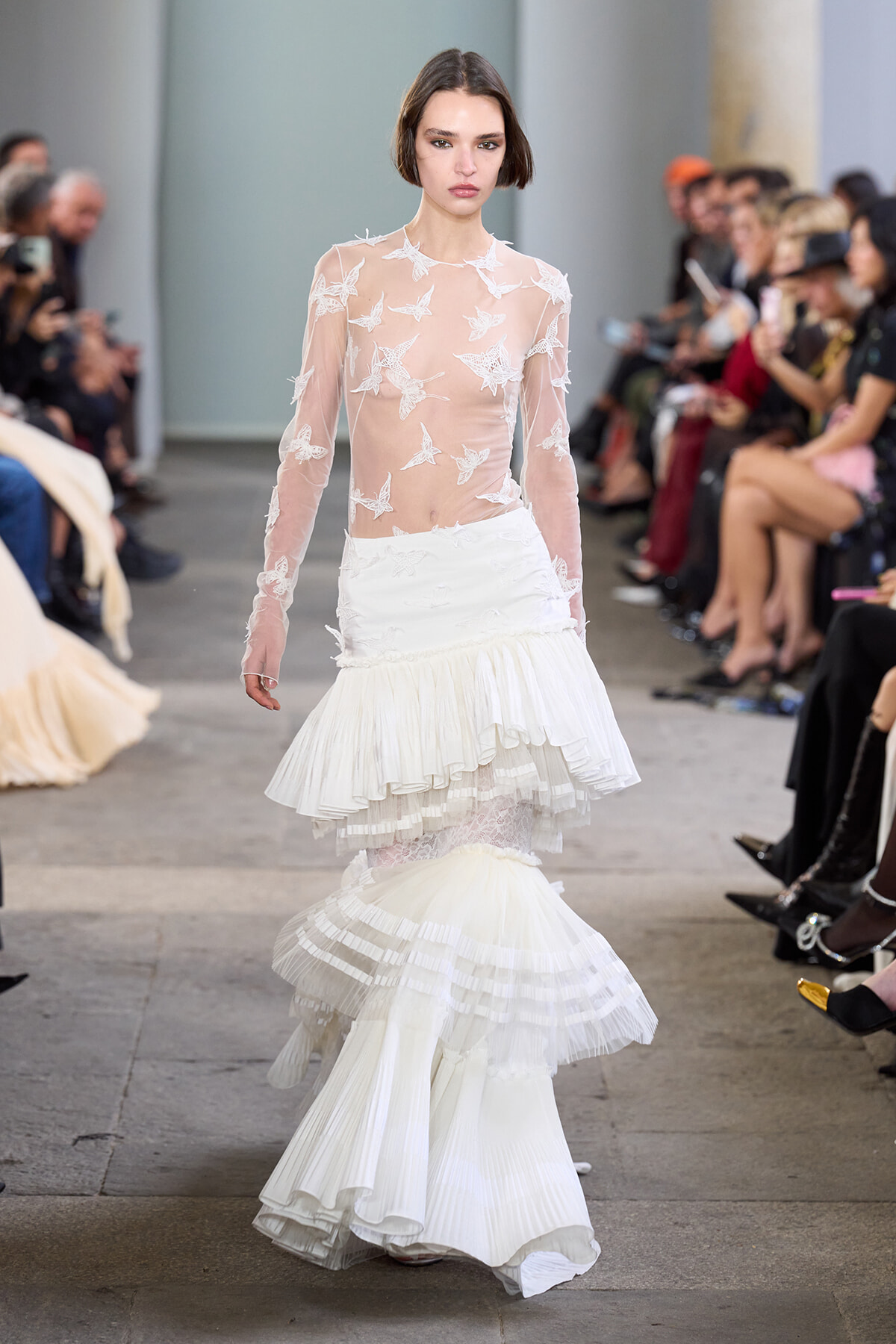 Model walks the runway in a sheer white lace top with star appliqués and a multi‑tiered white ruffled skirt on the catwalk.