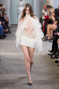 Model walks the runway in a sheer lace blouse with butterfly appliqués and a white mini skirt, paired with black heels; audience blurred in the background.