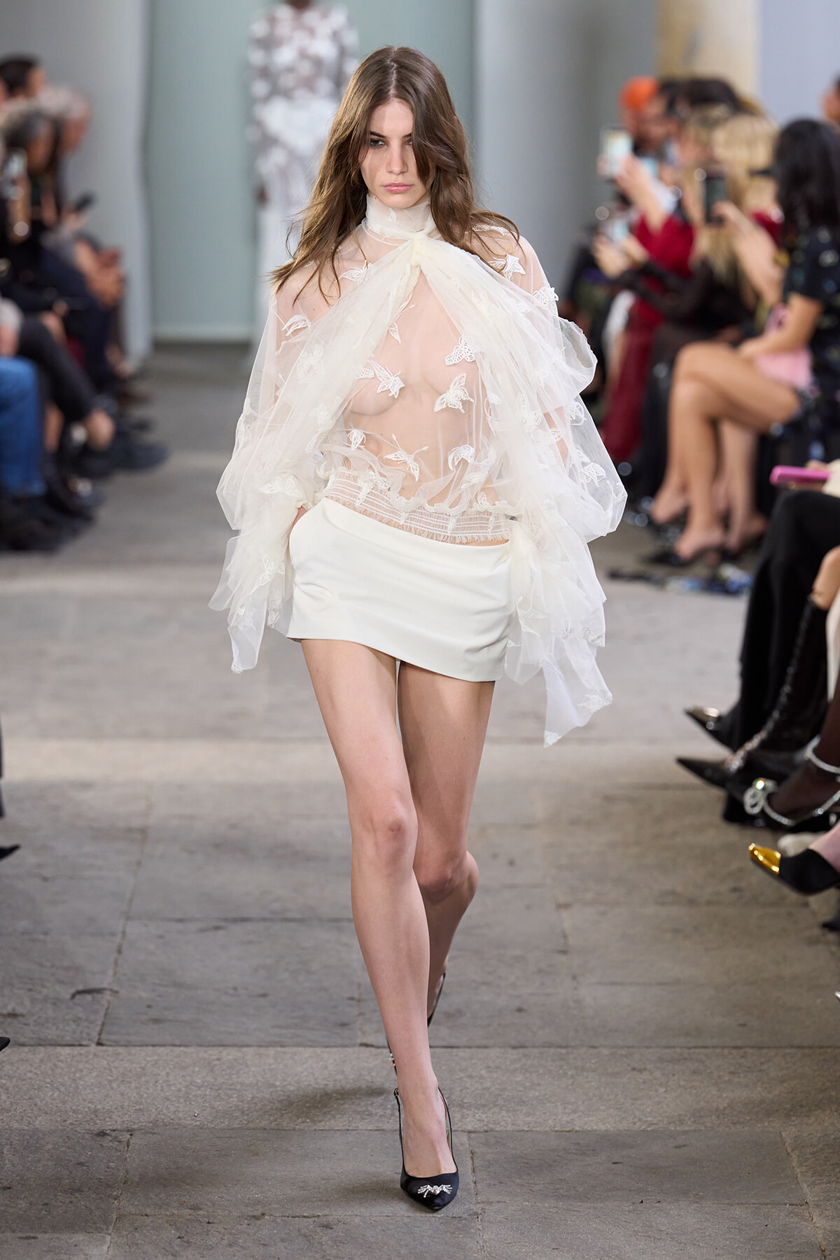 Model walks the runway in a sheer lace blouse with butterfly appliqués and a white mini skirt, paired with black heels; audience blurred in the background.