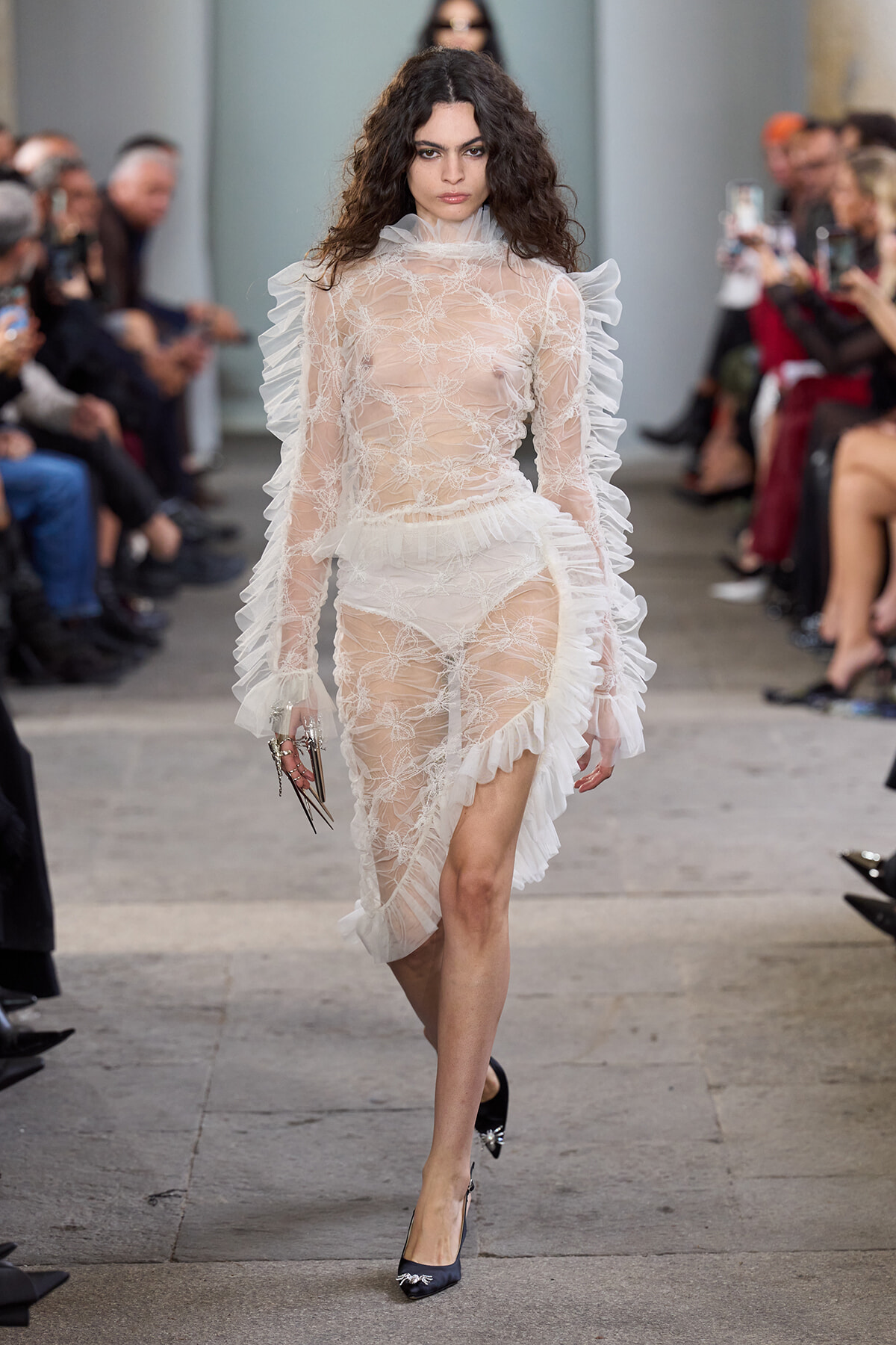 Model on a runway wearing a sheer white lace gown with ruffled tulle sleeves and high neckline, audience visible along the sides.