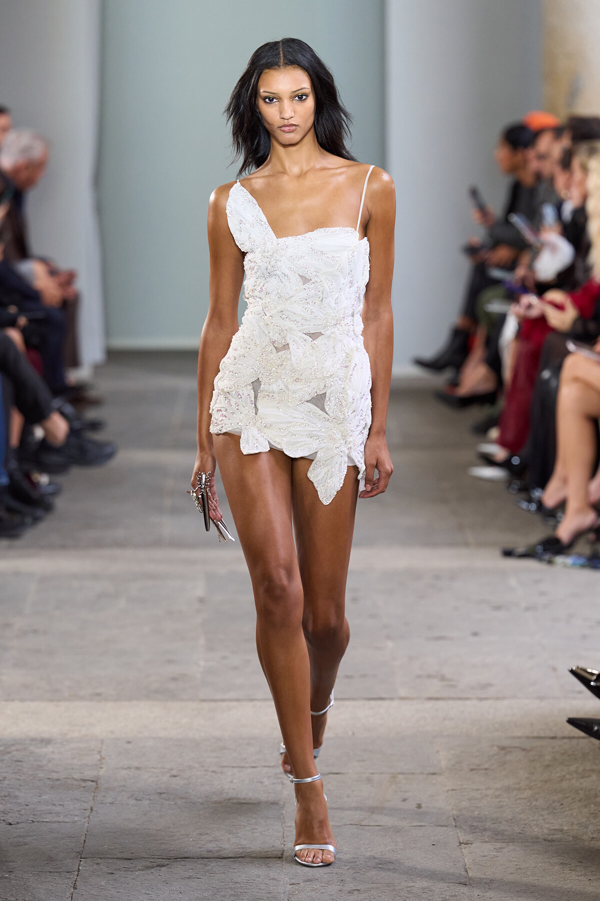 Model walks the runway in a white, lacy mini dress with floral appliqué and a single thin strap.