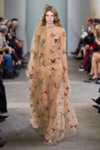 Model walks the runway in a sheer nude gown adorned with colorful butterfly embroidery.