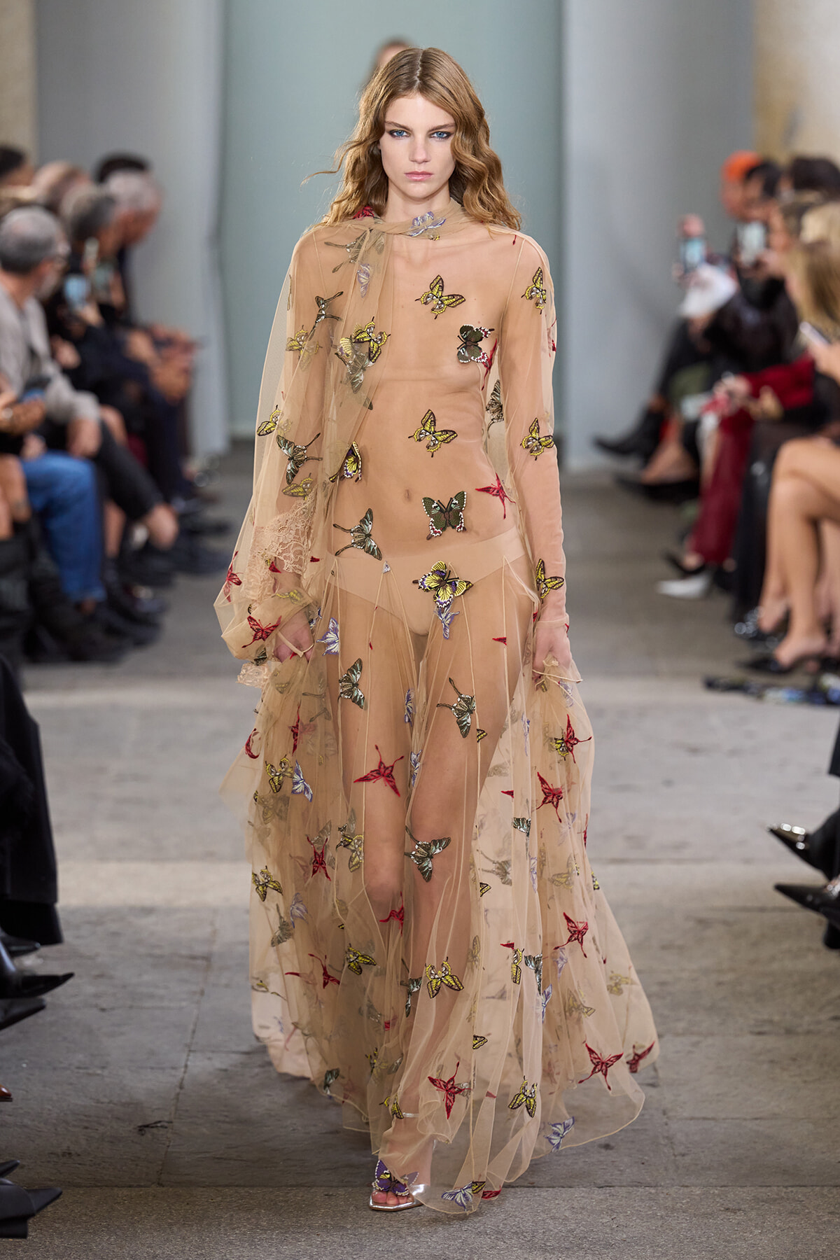 Model walks the runway in a sheer nude gown adorned with colorful butterfly embroidery.