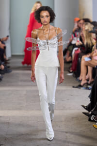Model walks a runway in a white sleeveless peplum top and white pants, with a large metallic dragonfly accessory across the chest; audience seated along the catwalk.