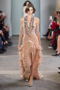 Model in a peach strapless dress with layered ruffles and long silver necklaces walks the runway.