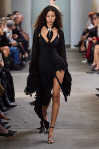 Model struts down a runway in a black, ruffled dress with a plunging neckline and sheer sleeves, wearing a silver necklace and strappy heels.