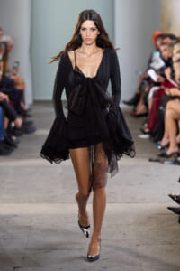 Runway model in a black dress with a deep V-neck and large lace bow at the waist, with sheer lace overlay on one leg and lace-trimmed sleeves.