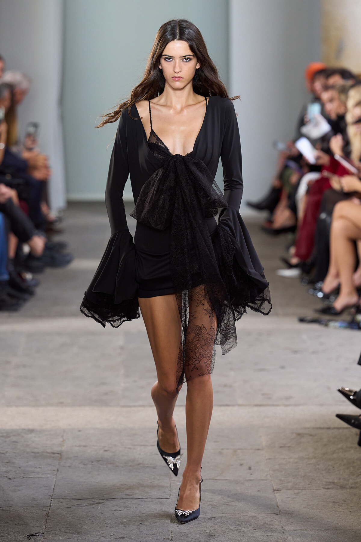 Runway model in a black dress with a deep V-neck and large lace bow at the waist, with sheer lace overlay on one leg and lace-trimmed sleeves.