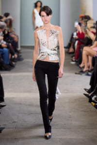 Model walks the runway in a printed, sleeveless wrap-top and black slim pants, with seated audience along the catwalk.