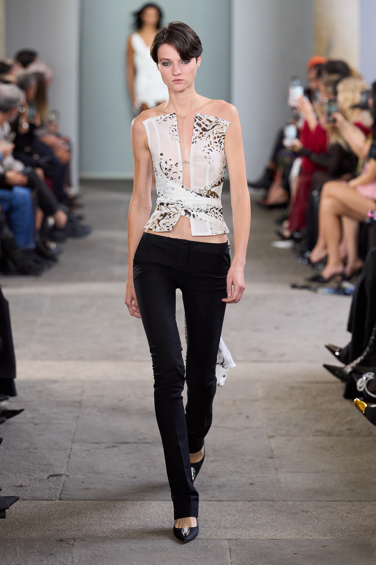 Model walks the runway in a printed, sleeveless wrap-top and black slim pants, with seated audience along the catwalk.