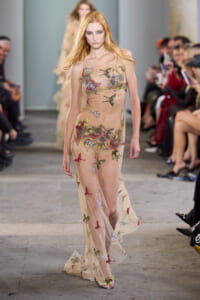 Female model walking the runway in a sheer beige gown covered with colorful butterfly embroidery, audience on both sides of the catwalk.