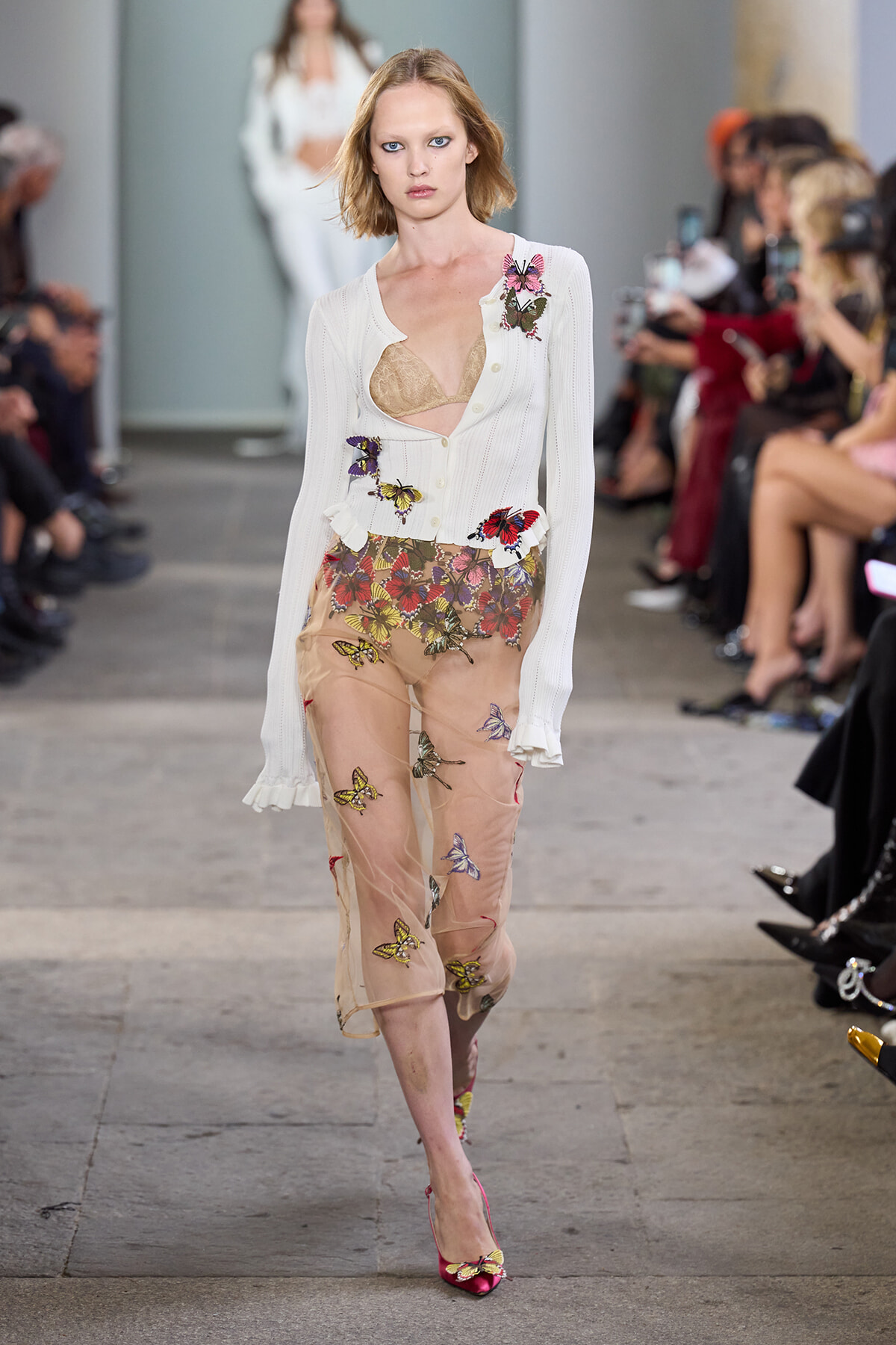 Model walking a runway in a white cardigan with butterfly embroidery over a sheer peach skirt; pink heels visible.