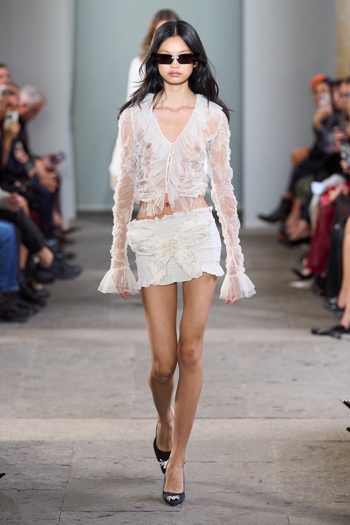 Model walks a runway in a sheer white lace blouse and short skirt with a large butterfly appliqué, sunglasses.